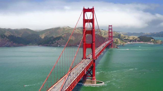 Vibrant aerial 4K footage of the beautiful suspension bridge known as Golden Gate Bridge against the backdrop of the hilly coast and cloudy sky. The water of the strait has turquoise color.