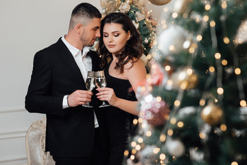 A bearded guy with a girl pose in a studio. Loving couple, is stylishly dressed near a Christmas tree. Christmas time. Christmas mood. Loving couple celebrating New Year and Christmas with champagne. 