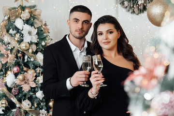 Beautiful couple holding champagne glasses. Lovely couple near Christmas tree. Newlyweds pose in a studio with an interesting light. A bearded guy with a girl is celebrating Christmas