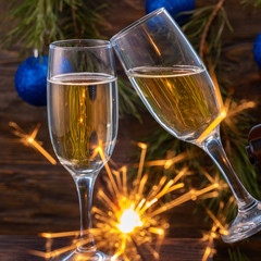 Glasses of champagne with bubbles on the background of Christmas decorations. Glasses touch during a festive toast and sparklers burn beautifully. Beautiful card. Square.