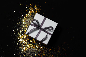 White gift box with silver ribbon and gold confetti and sparkles on black background. Top view, flat lay.