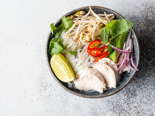 Traditional Vietnamese soup- pho ga in bowl with chicken and rice noodles, mint and cilantro, red onion, chili, bean sprouts and lime on grey background. Asian food.