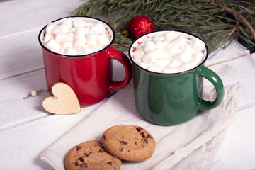 two mugs red and green with cocoa and marshmallows, homemade cookies on a napkin, spruce branches on a light background, top view