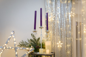 Purple candles in candlestick, fir tree branches and cones, siver curtains with shiny stars and warm garland lights and bokeh. 