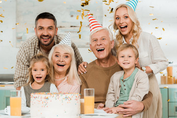 happy family looking at camera while sitting near birthday cake under falling confetti