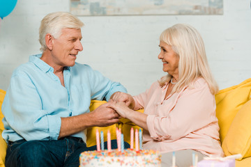 happy senior husband and wife sitting near birthday cake, looking at each other and holding hands