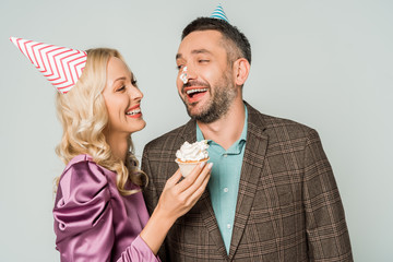 cheerful woman holding birthday cupcake near smiling husband with whipped cream on nose isolated on grey