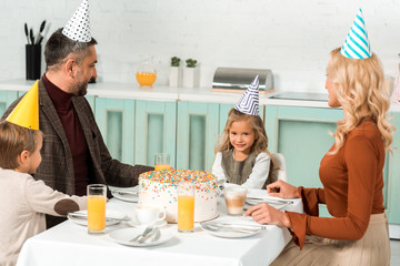 cute kid looking at birthday cake while sitting at served table near parents and brother