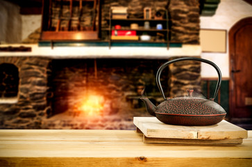 Metal kettle on wooden table.Free space for your decoration.Blurred background of fireplace with orange fire. Cold december night in home interior.Hot drink of tea,coffee or mulled.