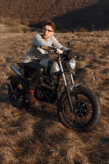Plakat Young male in eyeglasses and gloves motorcyclist travels on motorbike, treveler in desert, looking at a side in sunset time.