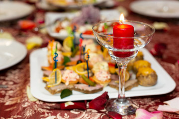 Beautifully laid table for the holiday. A candle in a glass goblet next to a plate of food appetizers on a red tablecloth in restaurant