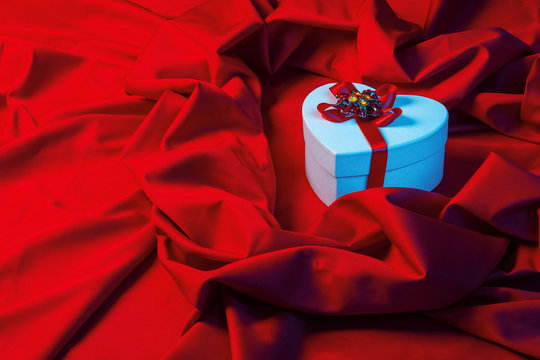 valentines greeting card in blue light. white cardboard box in shape of heart wrapped in ribbon lay on a red cloth which repeats the form of present package. love and romance gift concept
