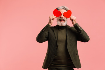 Happy bearded and mustache mature man in khaki suit holding two red hearts near eyes greeting...