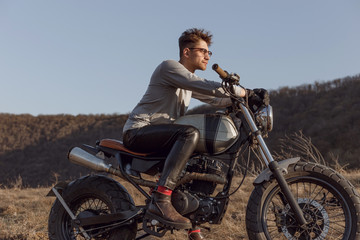 Good looking biker man wearing leather pants, helmet and eyeglasses sitting on his vintage...