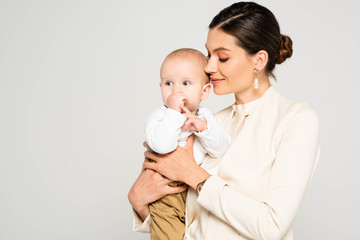 attractive positive businesswoman holding baby boy on hands, isolated on grey