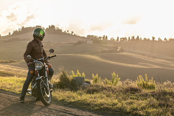 Man on his motorbike standing on a country road on hills landscape at sunset. Tuscany, Italy.
