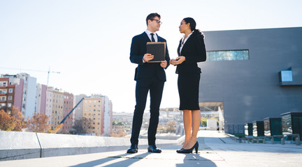 Formal young coworkers talking on street Modern multiracial woman and man with clipboard standing on street with modern architecture and having professional conversation in back lit