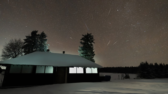 Night sky filled with stars including multiple Geminids meteors.  An old cabin is in the foreground set in a snowy scene and with lights on inside the cabin.  Pine trees as silhouettes. 