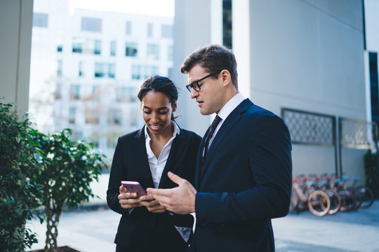 Modern young colleagues sharing smartphone Smiling ethnic businesswoman with diverse male coworker surfing mobile phone while chatting and standing on background of modern city