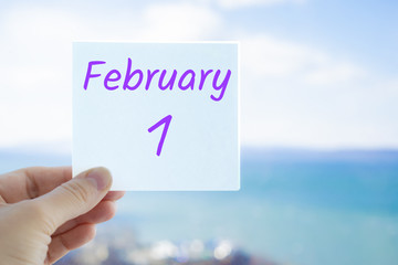 February 1st. Hand holding sticker with text February 1 on the blurred background of the sea and sky. Copy space for text. Month in calendar concept