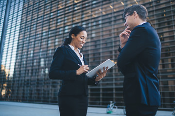 Cheerful woman using tablet while surveying man Elegant ethnic businesswoman in suit working on...