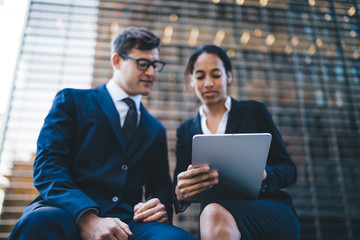 Diverse colleagues sharing tablet on street Low angle of modern young multiethnic woman and man sitting together on bench and using tablet against modern building exterior