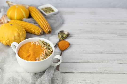 pumpkin cream soup in a ceramic plate on a white wooden background using gray textile, and small decorative pumpkin and corn and spoons. copy space