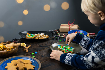 The child decorates with candys a gingerbread in the shape of a Christmas tree. Close up children's hands, lots of sweets on a wooden table.