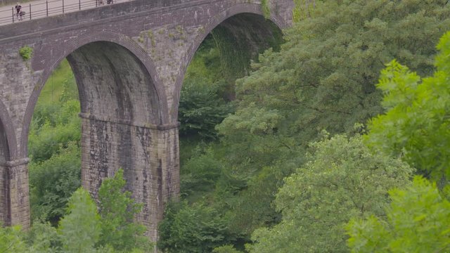 Medium low angle still shot of arched Monsal Headstone Viaduct Bridge with thick lush green valley vegetation, and cyclists riding at the top, Monsal Trail, Peak District, UK