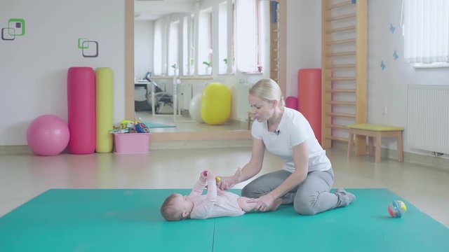 Woman teaching baby how to turn on stomach. Wide shot of a physiotherapist gym with person in focus sit on floor.