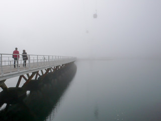pedestrians crossing a long wooden bridge over calm water on a foggy and misty morning