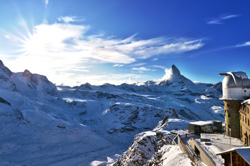 View of the Matterhorn, and snow-capped mountains. in the foreground the hotel and the observatory. In the background the blue sky with some clouds.