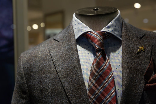 A dark gray jacket, a tie with blue and Burgundy stripes, a blue shirt with a pattern of squares hang on a mannequin behind glass in the window of a fashionable men's clothing store.