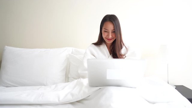 medium locked shot of an adult Asian woman working on a laptop in bed, smiling pose. All white and modern designed shot. Remote online working at home concept