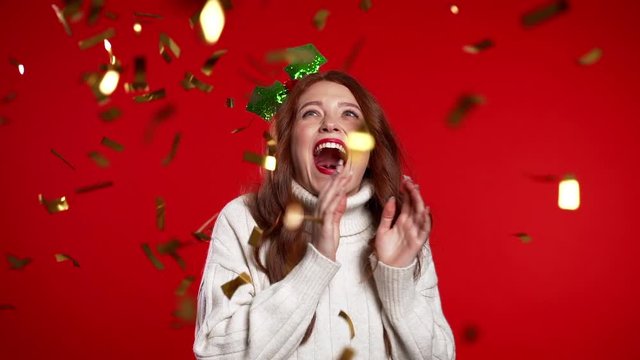 Excited woman in mistletoe head wreath dancing, applauding, having fun, rejoices over confetti rain in red studio. Concept of Christmas, New Year, happiness, party, winning.