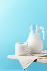 Glass jug and mug with milk on the edge of a white wooden on a blue background.