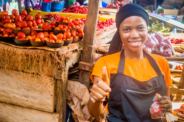 young african woman selling tomatoes in a local african market smiling an giving thumbs up gesture