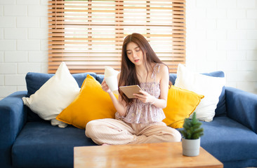 Portrait of young beautiful Asian woman at home sitting on the couch using tablet and drinking cup of hot coffee or tea in her living room. Lifestyle, Relaxation, Communication and technology concept.