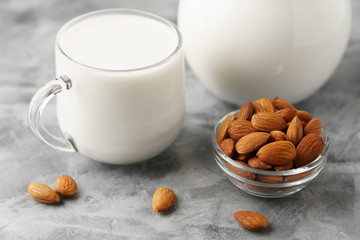 Almond milk in a glass jug and mug, next to nuts in a bowl on a concrete background.