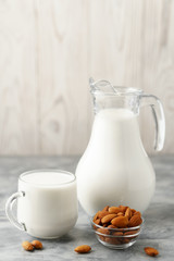 Almond milk in a glass jug and mug, next to nuts in a bowl on a concrete background.
