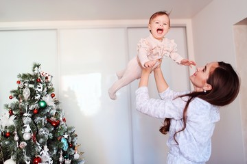 Happy beautiful woman having fun with her little daughter near the Christmas tree. Mom holds a cheerful baby in her arms