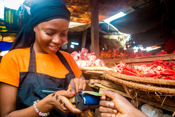 young african woman selling tomatoes in a local african market using her mobile pos device to...