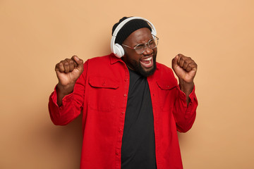 Overjoyed black bearded man raises clenched fists, sings song aloud, dances and enjoys music, wears white stereo headphones, red shirt, isolated over brown background, cheers indoor. Nice melody