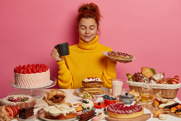 Pretty ginger female in yellow sweater, holds plate with delicious homemade pie, drinks tea, sits...