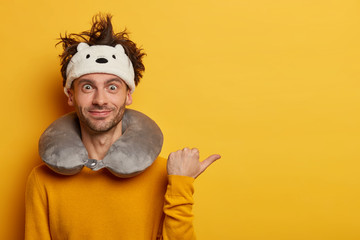 Glad European man points thumb away, dressed in yellow casual jumper, demonstrates blank space, wears travel pillow, travels in plane. People, traveling lifestyle, departure, voyage concept.
