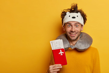 Cheerful man tourist waits for plane, holds passport and tickets, wears neck pillow, being in good mood, looks positively at camera, enjoys comfortable journey, isolated on yellow background.