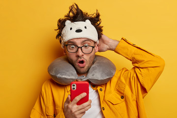 Tourist man stares at modern electronic device, keeps mouth opened, wears travel pillow not to have stiffness in transport, eye mask for sleeping, has business trip, isolated over yellow background
