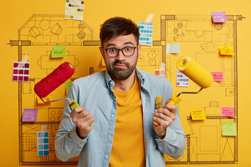 Doubtful bearded painter man paints walls with roller, chooses between red and yellow color, busy...