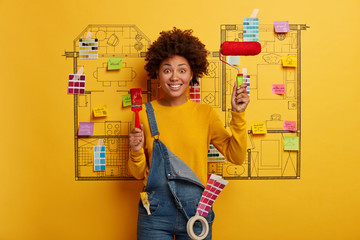 Pleased female holds repairing tools, uses paintbrush and roller for decorating room, has happy mood, implements modern interior, dressed in yellow jumper and denim overalls, improves house.