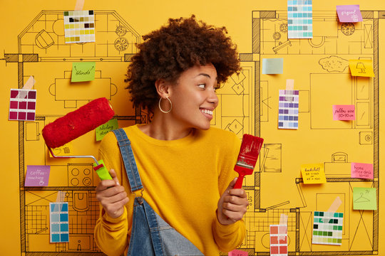 Horizontal view of curly haired African American female decorator holds paint brush and roller, turns gaze away, thinks about design of home, busy repairing apartment, wears jumper and denim overalls